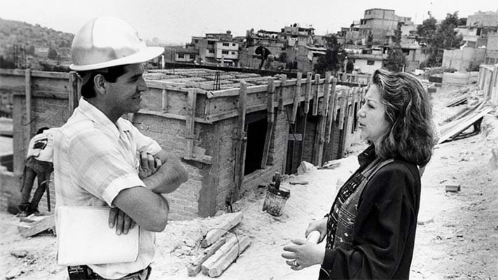 A black and white photo of a person wearing a construction hard hat and a person with shoulder length wavy hair talk to each other at a construction site.